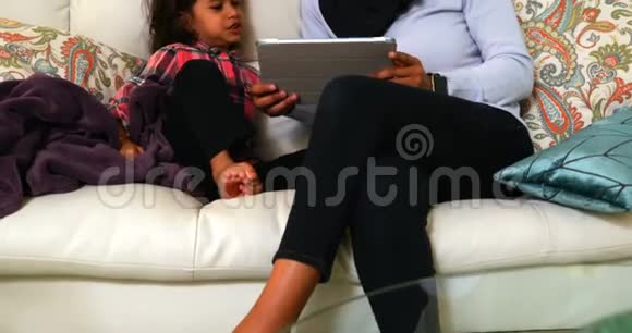 年轻的母亲在家里的沙发上和女儿一起使用数码平板电脑视频的预览图