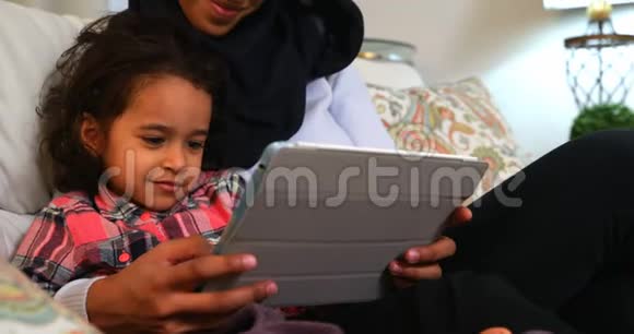 年轻的母亲在家里的沙发上和女儿一起使用数码平板电脑视频的预览图