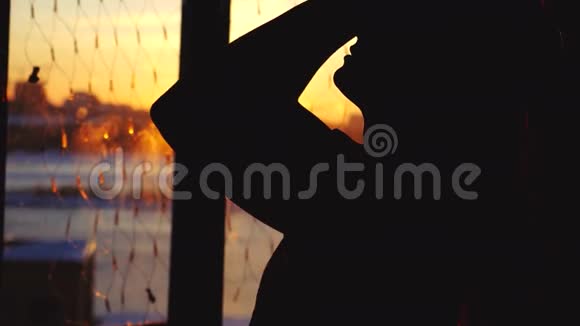 年轻女性的剪影在日落时在家中使用虚拟现实眼镜从窗户到城市背景视频的预览图