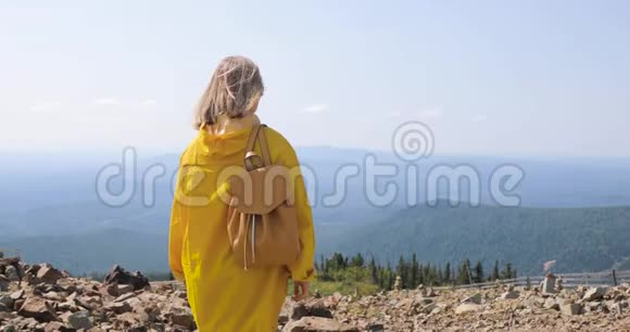 徒步旅行者在山顶上背包徒步旅行穿着黄色雨衣的女孩在山顶上视频的预览图