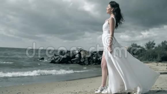 穿着白色连衣裙的漂亮女孩站在海滨着装随风飘扬慢动作视频的预览图