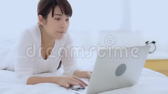 美丽的亚洲年轻女子微笑着躺在床上在卧室里用笔记本电脑休闲视频的预览图