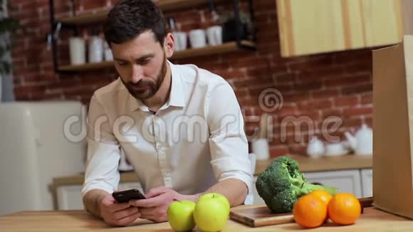 男人在家厨房用手机浏览英俊的年轻人在智能手机上浏览微笑着快乐视频的预览图