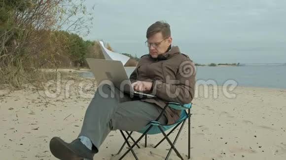 成熟的商人坐在河边放松穿暖和夹克用笔记本电脑的人视频的预览图
