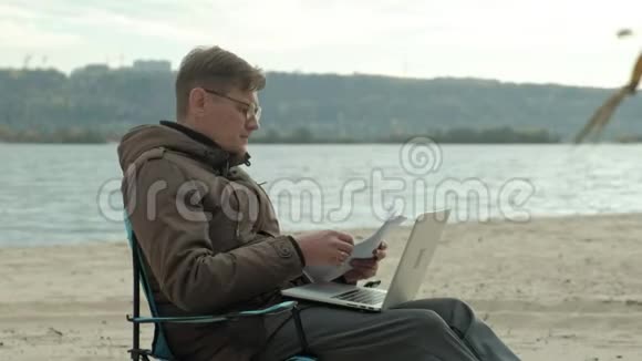 成熟的商人坐在河边放松穿暖和夹克用笔记本电脑的人视频的预览图