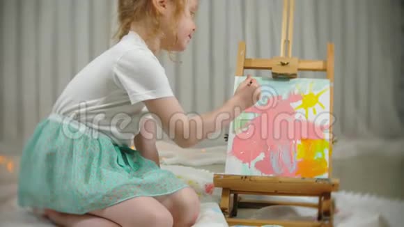 漂亮的红发女孩坐在画架前面的画室的地板上画画笔和彩色颜料视频的预览图