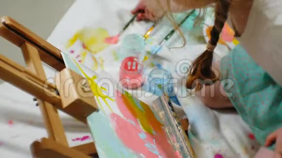 漂亮的红发女孩坐在画架前面的画室的地板上画画笔和彩色颜料视频的预览图