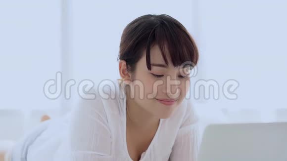 美丽的亚洲年轻女子微笑着躺在床上在卧室里用笔记本电脑休闲视频的预览图