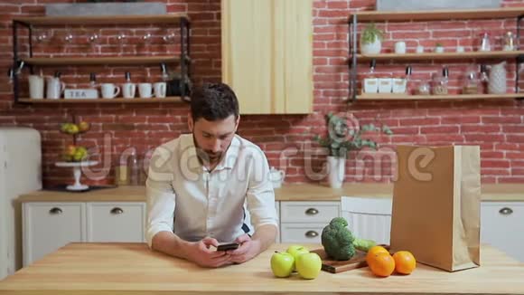 男人在家厨房用手机浏览英俊的年轻人在智能手机上浏览微笑着快乐视频的预览图