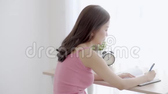 美丽的年轻亚洲女人微笑着坐在家里的客厅里学习写笔记本和日记视频的预览图