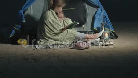 妈妈和她美丽的女儿晚上在海滩上的旅游帐篷附近读了一本书视频的预览图