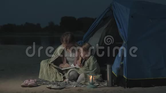 妈妈和她美丽的女儿晚上在海滩上的旅游帐篷附近读了一本书视频的预览图