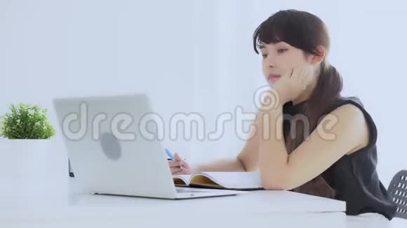 美丽的亚洲女人微笑着坐在客厅里学习在家写笔记本视频的预览图