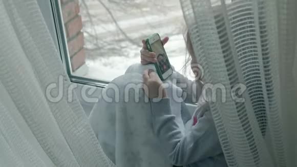 快乐的小女孩一头波浪状的红色头发坐在窗台上盖着毯子用电话说话视频视频的预览图