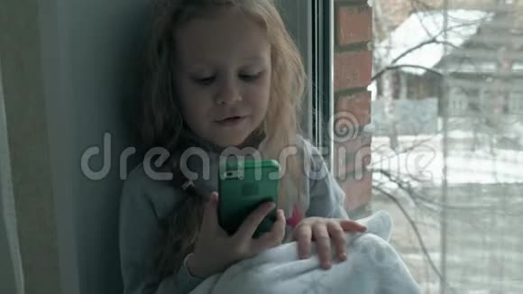 快乐的小女孩一头波浪状的红色头发坐在窗台上盖着毯子用电话说话视频视频的预览图