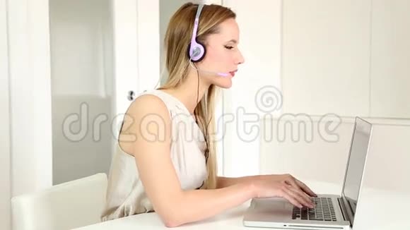 一位年轻漂亮的金发电话销售小姐她戴着耳机在电脑上通过电话与顾客交谈视频的预览图