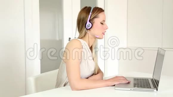 一位年轻漂亮的金发电话销售小姐她戴着耳机在电脑上通过电话与顾客交谈视频的预览图