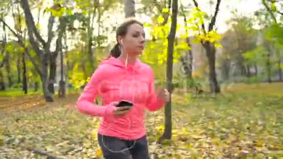 在日落时分一位戴着耳机和智能手机的女士在秋天的公园里奔跑视频的预览图