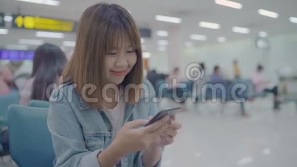 快乐的亚洲女人在机场登机口等待航班时在候机大厅使用和检查她的智能手机视频的预览图