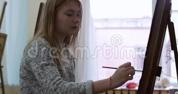 年轻美丽的女艺术家在一个艺术工作室坐在一个Easel后面和画布上的绘画绘图过程视频的预览图