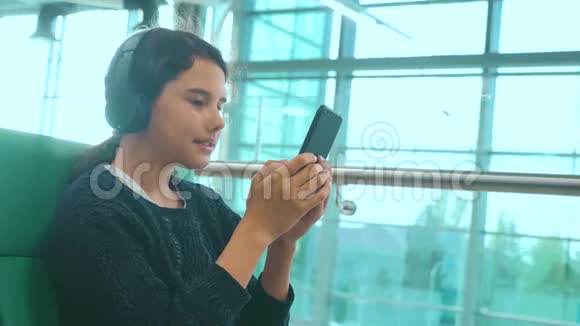 机场候机大厅供飞机飞行戴着耳机听音乐的年轻快乐少女视频的预览图