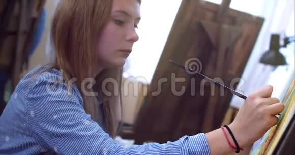 一位年轻美丽的女艺术家在一个艺术工作室坐在一个Easel后面和画布上的绘画绘图过程视频的预览图