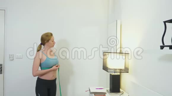 超重和减肥的概念一个女人在卧室里用卷尺测量自己在看着视频的预览图