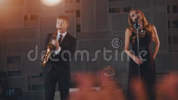 身穿黑色礼服的爵士歌手在台上与萨克斯演奏家一起在麦克风上表演视频的预览图