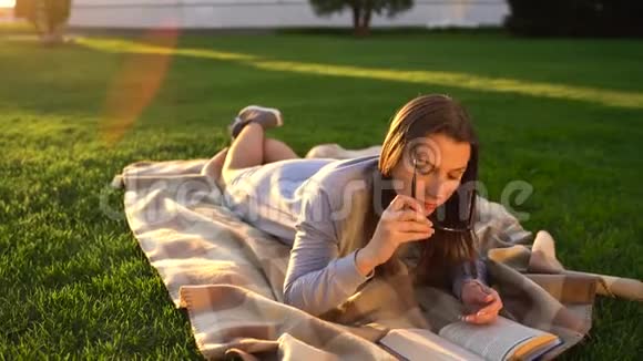 戴眼镜的女孩在日落时躺在公园的毯子上看书视频的预览图