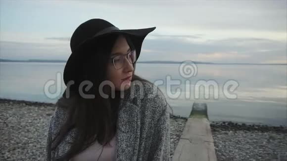 日落时分戴着黑色帽子和眼镜的年轻女子在海边视频的预览图