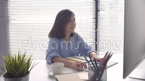 美丽的年轻微笑的亚洲女人在电脑上工作在家里的客厅喝咖啡视频的预览图