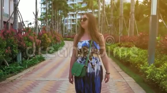 迷人的快乐女孩戴着眼镜和一件蓝色的裙子走在热带城市的街道上棕榈树和太阳视频的预览图