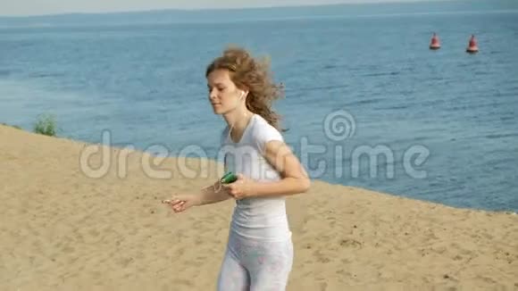 一个身材好的年轻女子在黎明时在海里从事体操她戴着耳机沿着海岸跑视频的预览图