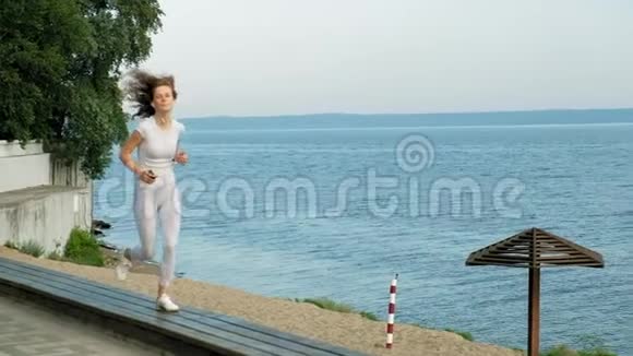 一个身材好的年轻女子在黎明时在海里从事体操她戴着耳机沿着海岸跑视频的预览图