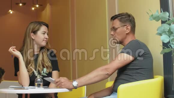 一个商务女性和一个男人在休息的时候拿着他们的平板电脑坐在一黄色墙壁的咖啡馆里讨论一个项目视频的预览图