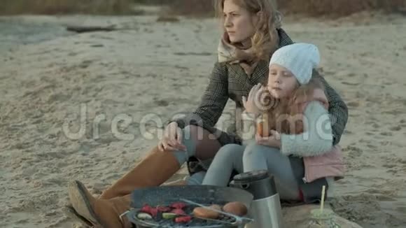 一个穿着外套的年轻女子和一个卷发的女孩一个母亲和一个女儿在河边的海滩上大海里野餐视频的预览图