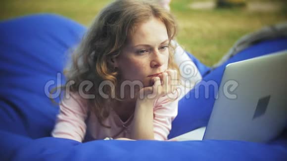 年轻女子用手提电脑躺在公园的一个休息区软泡芙上视频的预览图