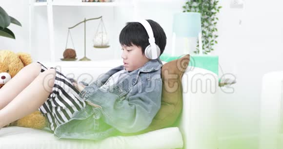 年轻的亚裔男孩在家玩带耳机的平板电脑视频的预览图