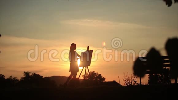 一个金发女孩的剪影借助颜料在画布上画一幅画一个木制画架保存着这幅画视频的预览图