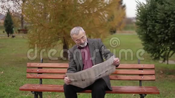 一位英俊的祖父穿着灰色夹克留着漂亮的胡子坐在公园的长凳上看报纸视频的预览图