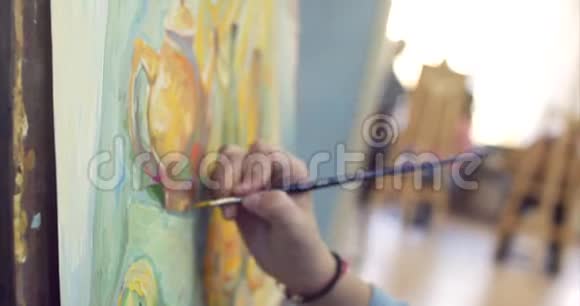 一位年轻美丽的女艺术家在一个艺术工作室坐在一个Easel后面和画布上的绘画绘图过程视频的预览图