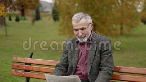 一位英俊的祖父穿着灰色夹克留着漂亮的胡子坐在公园的长凳上看报纸视频的预览图