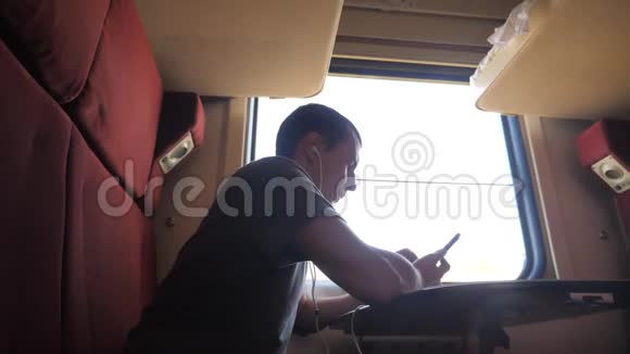 男子剪影旅行坐在火车车厢里坐在窗边一辆智能手机坐在铁路上戴着耳机视频的预览图