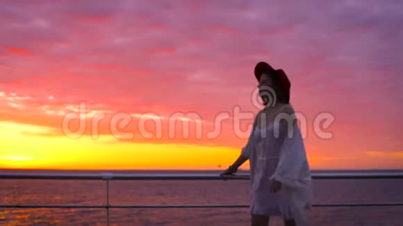 美丽的日落时分穿着白色连衣裙和戴着帽子的年轻迷人女子在海上背景下摆姿势视频的预览图
