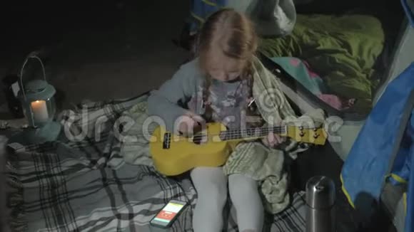 一个漂亮的女孩在一个旅游帐篷附近的河岸上的四弦琴上玩耍视频的预览图