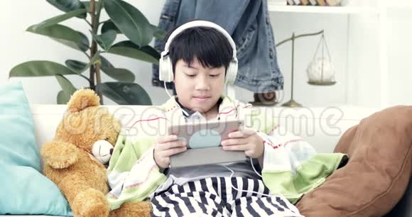 年轻的亚裔男孩在家玩带耳机的平板电脑视频的预览图