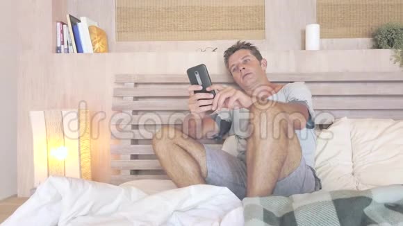 年轻有魅力的30岁男人蓝眼睛在家里躺在床上用手机上的网络社交媒体约会应用程序快乐放松视频的预览图