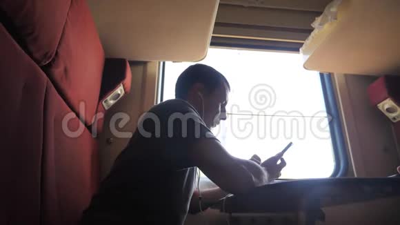 男子剪影旅行坐在火车车厢里坐在窗边一辆智能手机坐在铁路上戴着耳机视频的预览图
