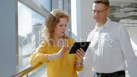 年轻漂亮的商业女人和成熟的男人靠窗拿着一块平板电脑视频的预览图