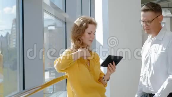 年轻漂亮的商业女人和成熟的男人靠窗拿着一块平板电脑视频的预览图